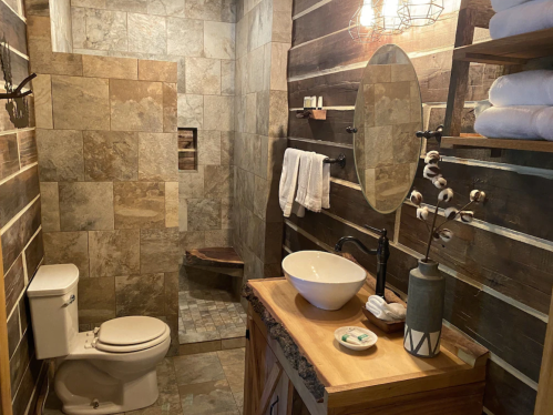 A rustic bathroom featuring stone tiles, a wooden vanity, a round mirror, and decorative towels and plants.