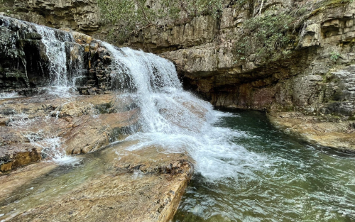 A serene waterfall cascading over rocky terrain into a clear pool, surrounded by lush greenery.