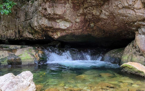 A serene stream flows under a rocky overhang, surrounded by mossy stones and clear water.