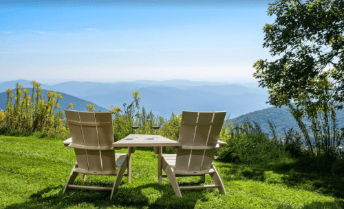 Two white chairs and a small table overlook a scenic mountain view, surrounded by greenery and flowers.