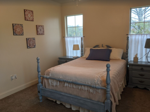 Cozy bedroom with a light blue bed, nightstands, and decorative wall art. Natural light from two windows.