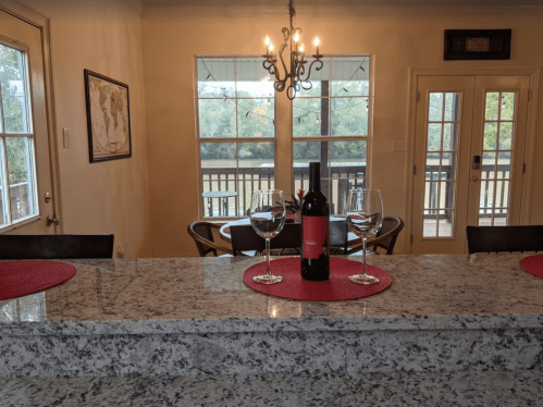 A cozy kitchen with a granite countertop, wine bottle, and glasses, overlooking a deck and scenic view through large windows.