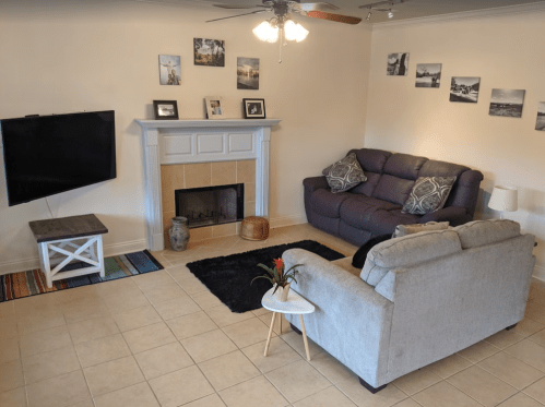 Cozy living room with a TV, fireplace, two sofas, and decorative photos on the wall. A rug adds color to the tiled floor.