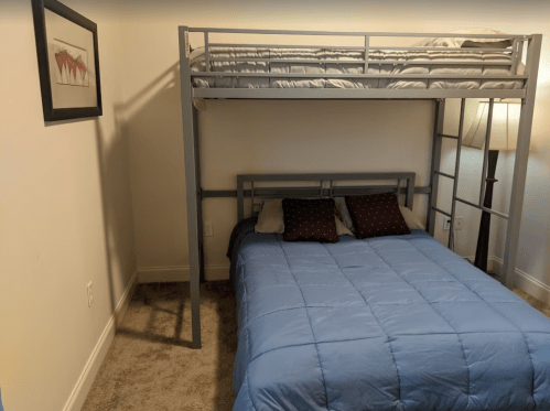 A room with a bunk bed and a double bed, featuring blue bedding and a lamp, with a framed picture on the wall.