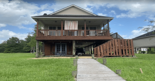 A two-story house with a wooden deck, surrounded by green grass and trees, under a partly cloudy sky.