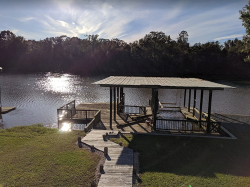 A serene riverside scene featuring a wooden dock and gazebo, surrounded by trees and reflecting sunlight on the water.