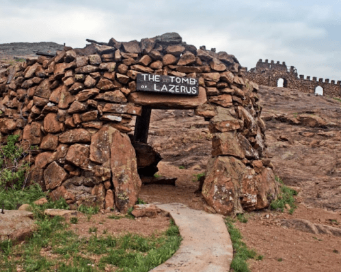 A stone structure with a sign reading "The Tomb of Lazarus," set against a rocky landscape under a cloudy sky.