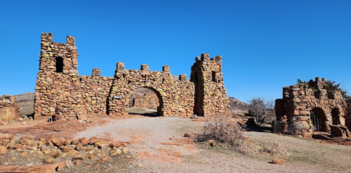 Stone ruins with towers and arches against a clear blue sky, surrounded by rocky terrain and sparse vegetation.