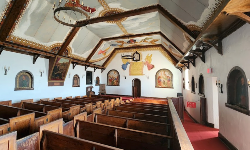 Interior of a church featuring wooden pews, colorful murals, and a high, decorated ceiling. Exit sign visible.