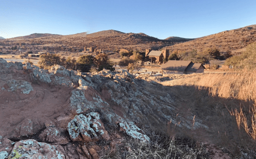A scenic landscape featuring rocky terrain, rolling hills, and rustic buildings under a clear blue sky.