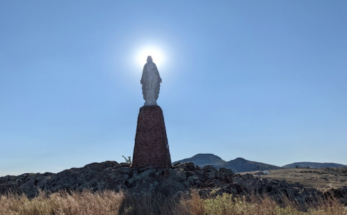 A statue of a figure stands on a rocky hill, silhouetted against the sun in a clear blue sky.