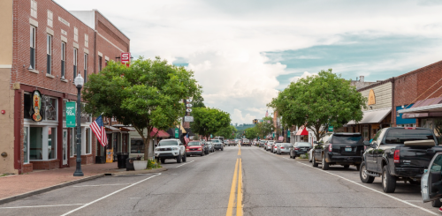 A quiet downtown street lined with shops and trees, featuring parked cars and a cloudy sky above.
