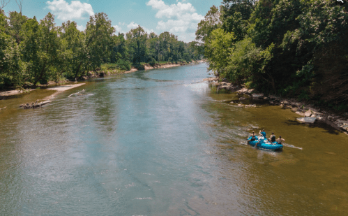 Two people in a blue raft float down a calm river surrounded by lush green trees and a clear blue sky.
