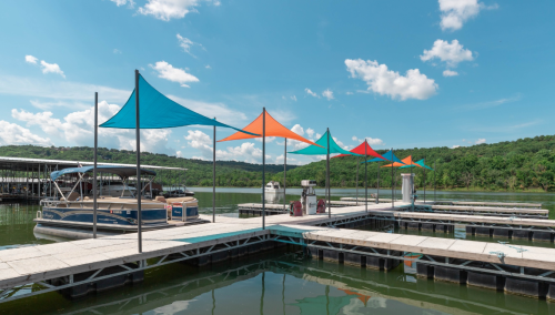 Colorful shade sails over a marina dock with boats, surrounded by green hills and a blue sky with fluffy clouds.