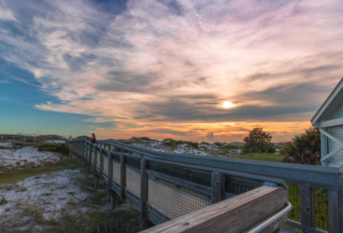 A wooden walkway leads to a beach at sunset, with colorful clouds and a hint of the sun on the horizon.