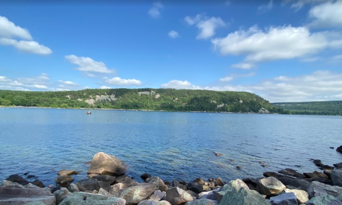 A serene lake surrounded by green hills and rocky shores under a blue sky with fluffy clouds.