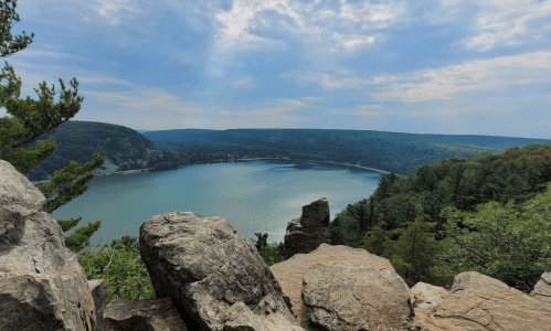 A scenic view of a lake surrounded by green hills and rocky terrain under a partly cloudy sky.