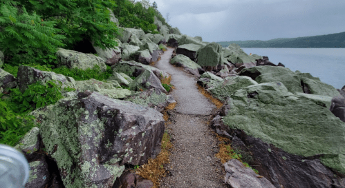 A rocky path lined with large stones, leading along a lake under a cloudy sky. Lush greenery is visible nearby.