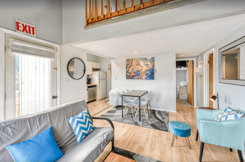 Cozy living space with a futon, dining table, and kitchen area, featuring light wood floors and colorful wall art.
