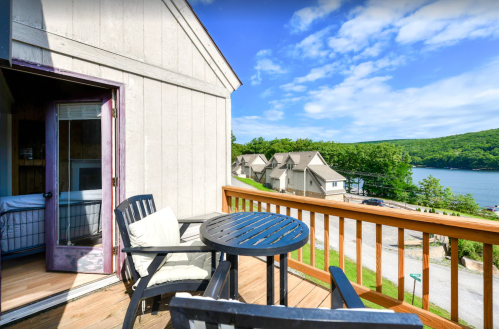 A balcony with a table and chairs overlooks a scenic lake and nearby cottages under a clear blue sky.