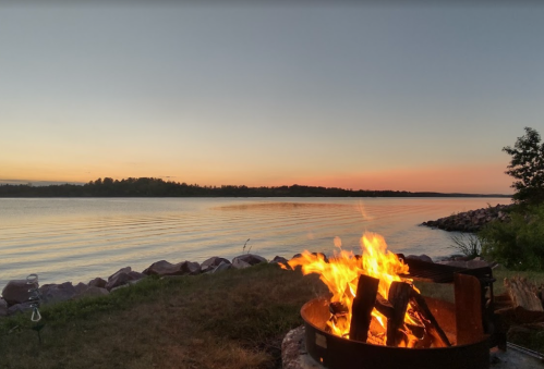 A cozy campfire burns by a calm lake at sunset, with trees and rocks in the background.