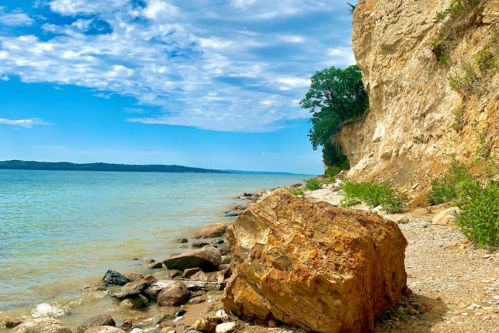 A rocky shoreline with a cliff, clear blue water, and a bright sky with scattered clouds.