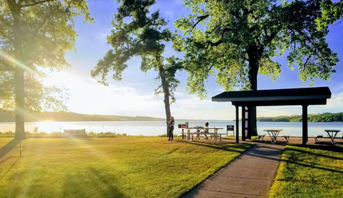A serene lakeside scene with picnic tables, trees, and people enjoying the sunset by the water.
