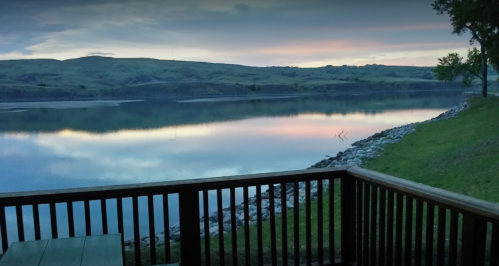 A serene lakeside view at dawn, with calm waters reflecting the colorful sky and green hills in the background.