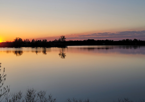 A serene sunset over a calm lake, with silhouettes of trees reflected in the water.