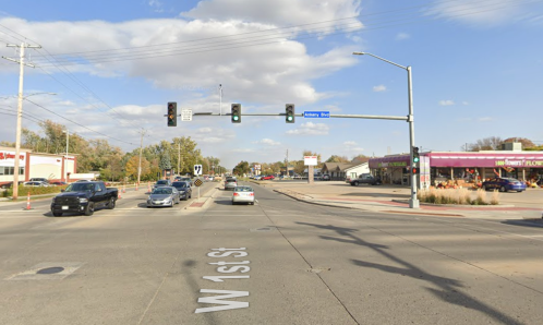 Traffic intersection with a green light, cars on the road, and businesses visible in the background under a cloudy sky.