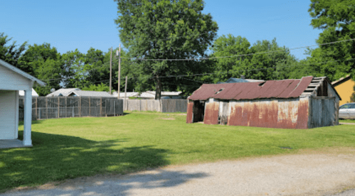 A grassy area with a rusty shed, wooden fence, and trees under a clear blue sky.