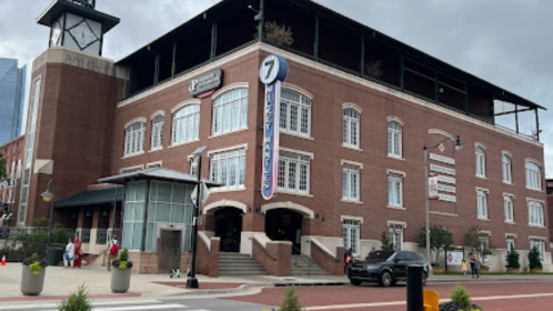 A brick building with large windows and a clock tower, featuring a sign for "7 Mile Market" and nearby pedestrians.