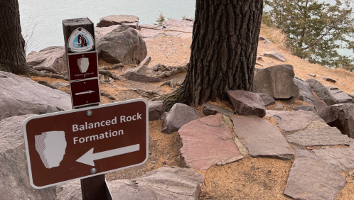 Sign for "Balanced Rock Formation" pointing left, with rocky terrain and trees in the background near a body of water.