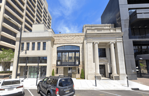 A street view of a historic building with columns, flanked by modern structures and parked cars under a blue sky.