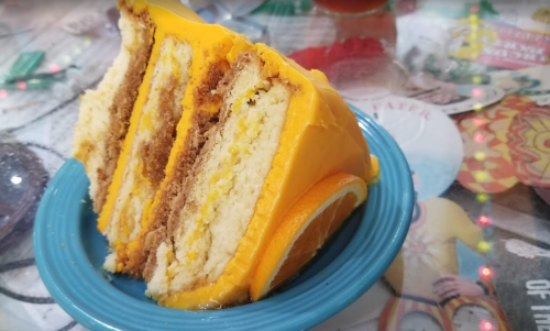 A slice of layered cake with orange frosting and a slice of orange on the side, served on a blue plate.