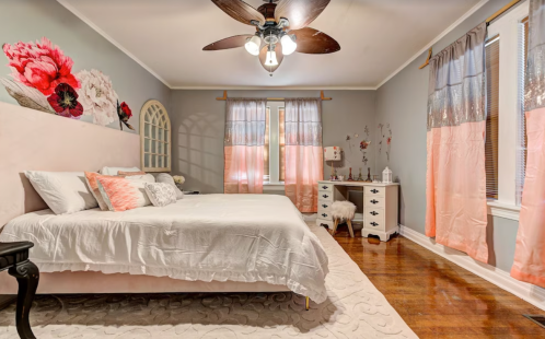 Cozy bedroom with a pink bed, floral wall art, wooden floors, and soft curtains, creating a warm, inviting atmosphere.