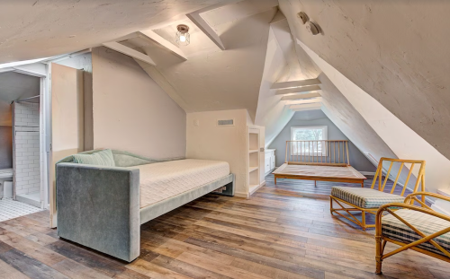 Cozy attic room with two beds, a chair, and wooden flooring, featuring sloped ceilings and natural light.