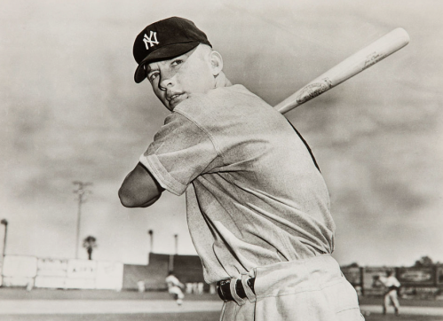 A young baseball player in a Yankees cap poses with a bat, ready to swing, against a cloudy sky backdrop.