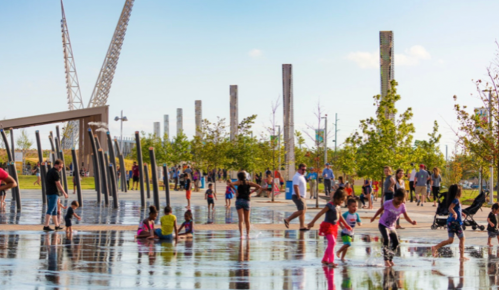 Children and families play in a splash pad area, surrounded by trees and tall pillars under a clear blue sky.