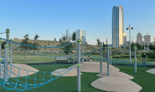 A playground with climbing nets and sand mounds, set against a city skyline and clear blue sky.
