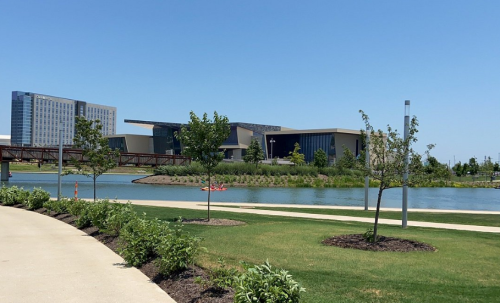 A scenic view of a park with a pathway, water, and modern buildings in the background under a clear blue sky.