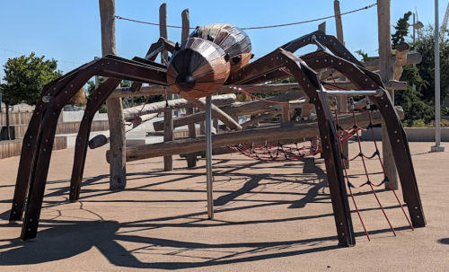 A large wooden spider playground structure with climbing features, set in a sunny outdoor area.