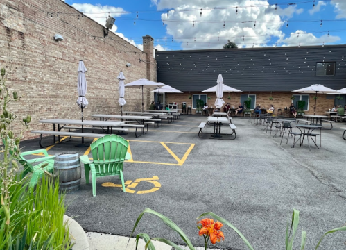 Outdoor seating area with picnic tables, umbrellas, and green chairs, surrounded by a brick wall and string lights.