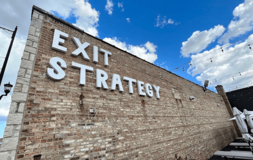 A brick wall with large white letters spelling "EXIT STRATEGY" against a blue sky with scattered clouds.