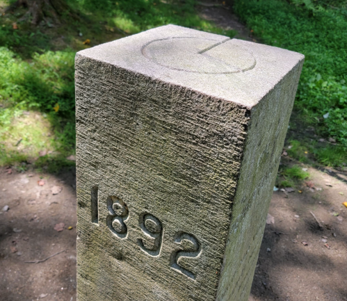 A weathered stone pillar marked with the year "1892" and a circular design on top, set in a grassy area.