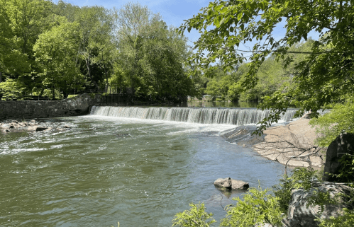 A serene river scene featuring a waterfall surrounded by lush green trees and rocky banks under a clear blue sky.