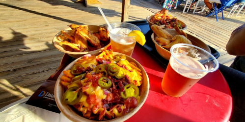 A table with three bowls of food, including loaded fries, and two drinks, one with a lemon slice, on a wooden deck.