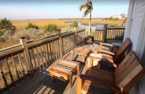 A wooden deck with colorful chairs and tables overlooks a marshy landscape and winding waterway.