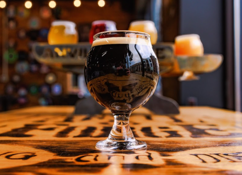 A dark beer in a glass sits on a wooden table, with various other beers blurred in the background.