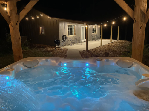A lit hot tub with blue water in the foreground, and a cozy cabin with string lights in the background at night.